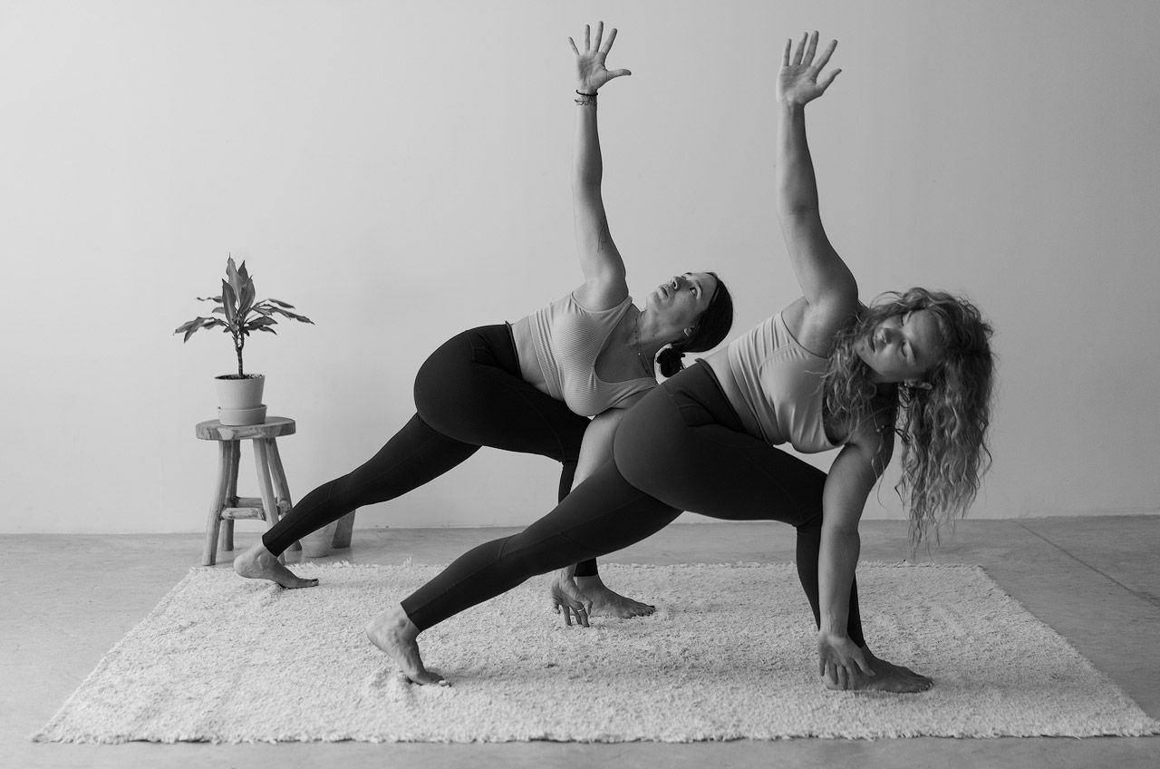 Dos mujeres en blanco y negro realizan la pose de yoga de Ángulo Lateral Extendido en Torsión (Parivrtta Parsvakonasana) sobre una alfombra blanca. Formación de profesora de yoga